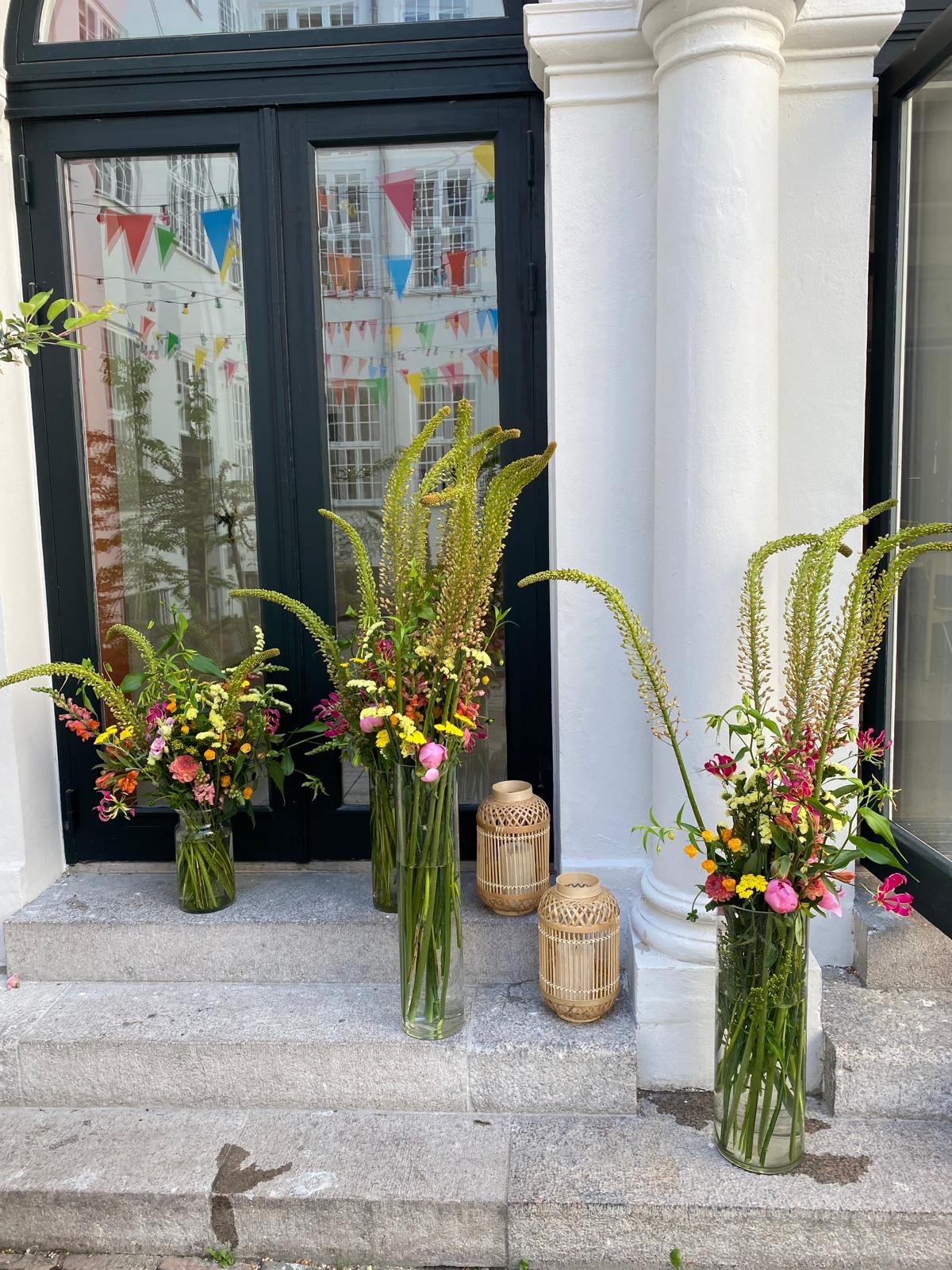 Tre høje glasvaser med blomsterspirer foran en entré